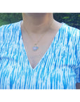 A woman wearing a blue and white patterned top with a gold opal pendant necklace outdoors.