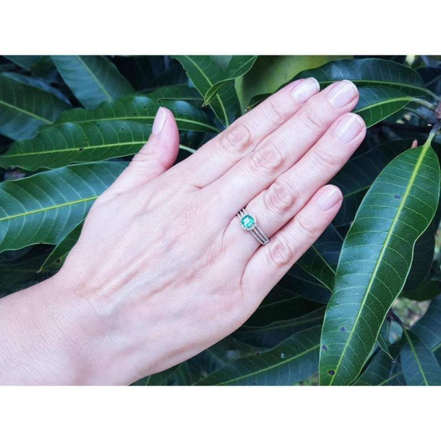 Hand wearing a 14K white gold emerald and halo diamond engagement ring against green leaves.