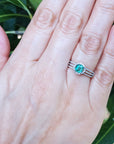 A natural emerald engagement ring in white gold on a person's hand against a green natural background.