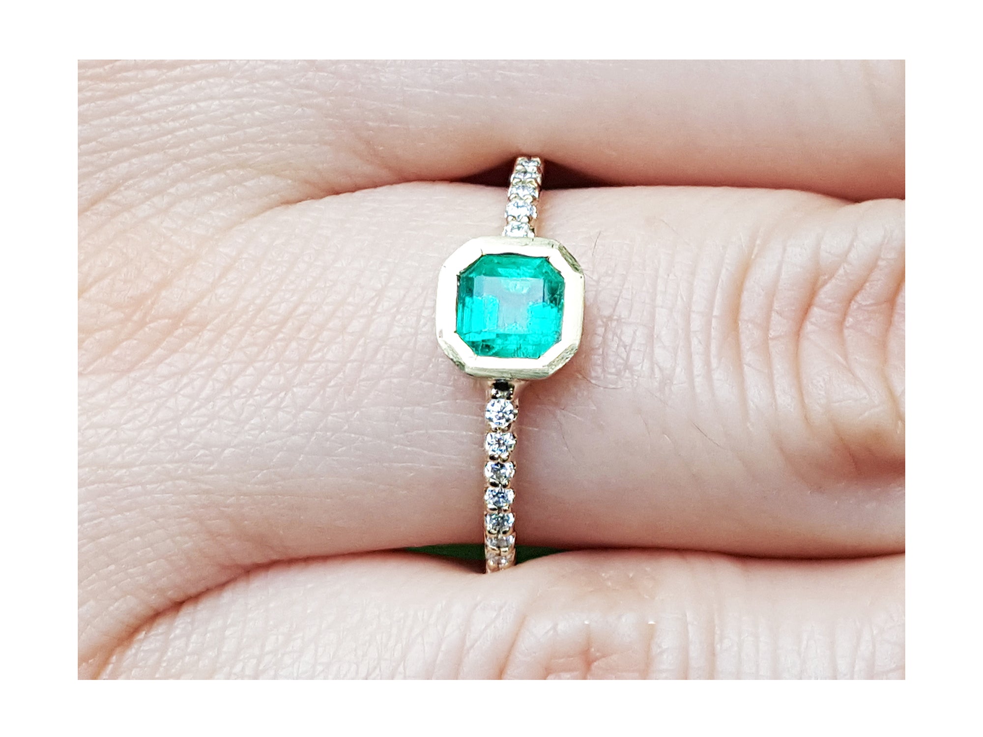 A yellow gold ring with a bezel set emerald on a finger.