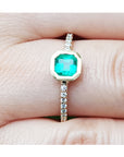 A yellow gold ring with a bezel set emerald on a finger.