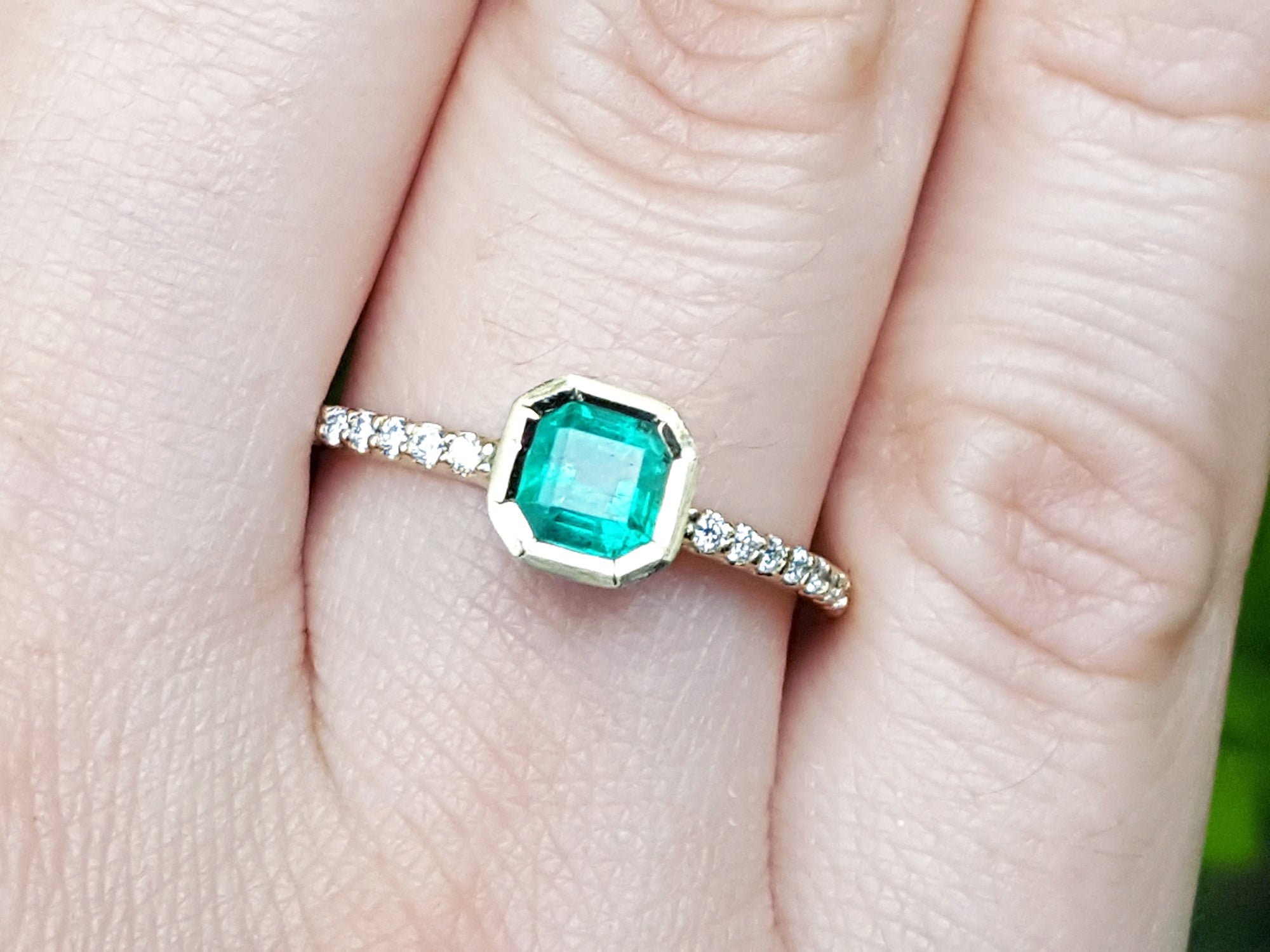 A 14k yellow gold emerald ring bezel set with accent diamonds on a finger.