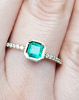 A 14k yellow gold emerald ring bezel set with accent diamonds on a finger.