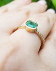 Gold ring with a natural emerald on a hand against a blurred green background.