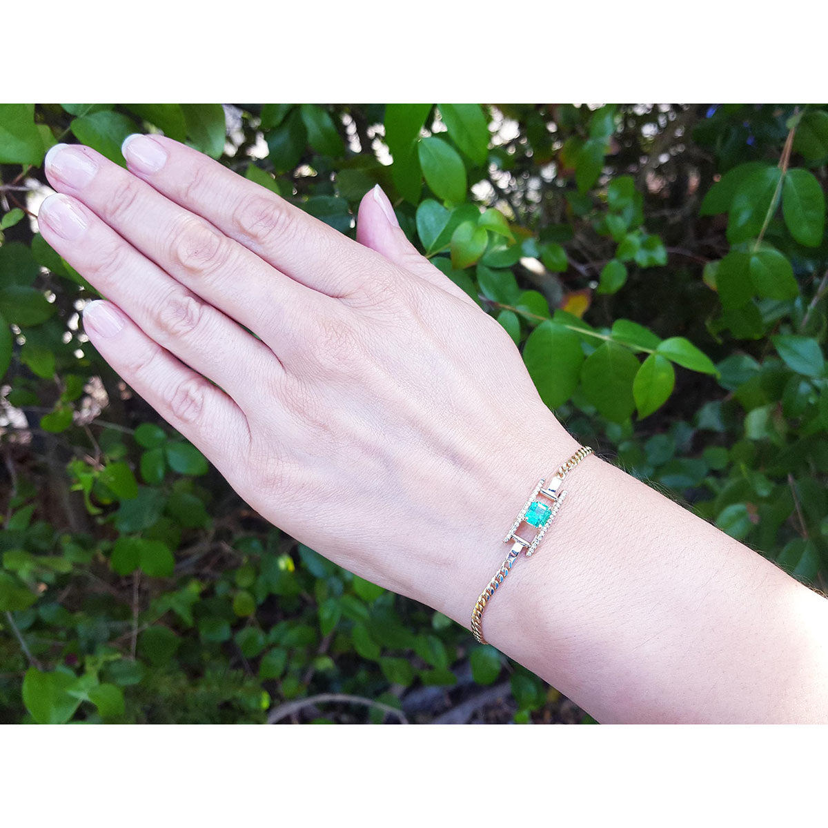 Hand wearing a bracelet with natural emerald against a green leafy background.