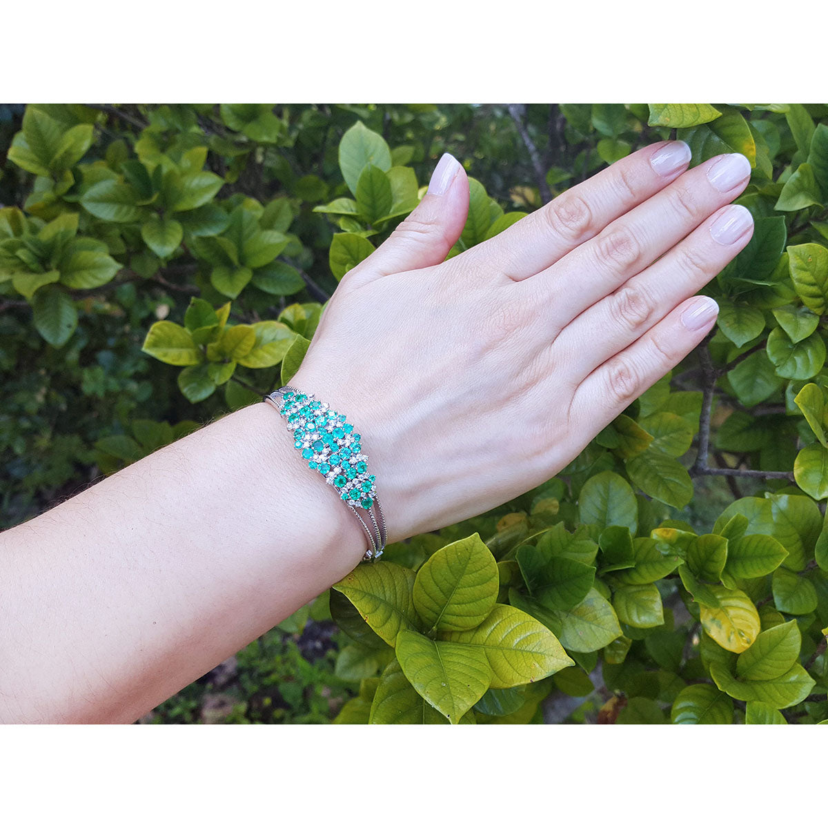 Hand wearing a white gold emerald bangle bracelet with a green leafy background.