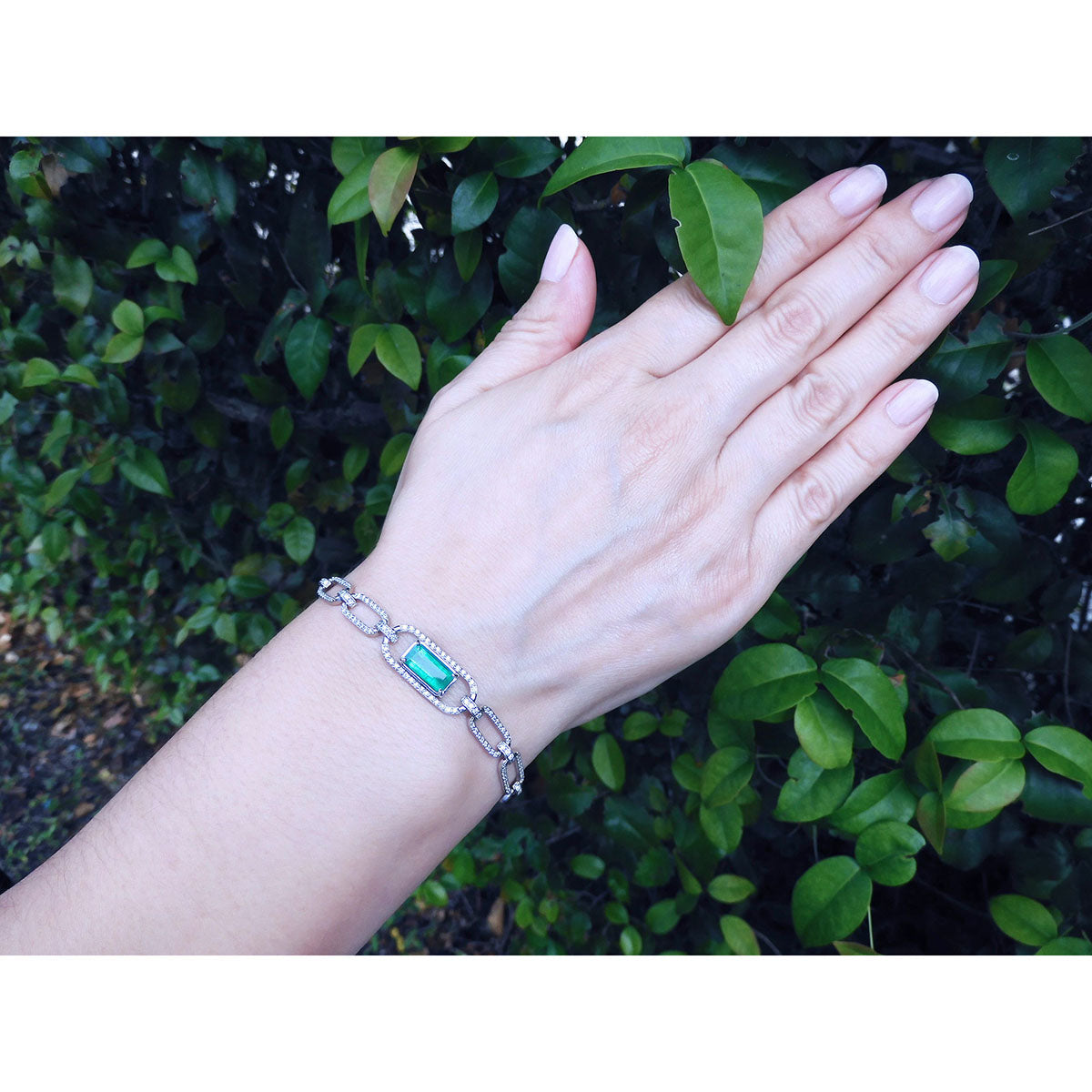 A woman&#39;s hand wearing a paperclip bracelet with a colombian emerald against a green leafy background.