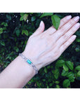 A woman's hand wearing a paperclip bracelet with a colombian emerald against a green leafy background.
