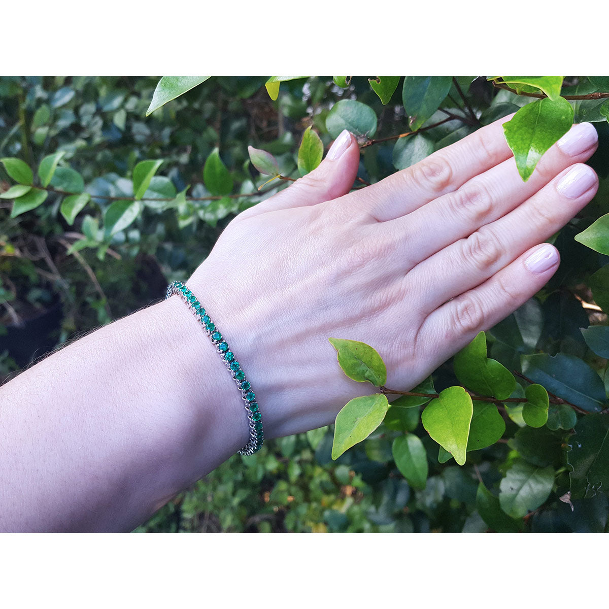 Hand wearing a natural round cut emerald tennis bracelet with a leafy background.