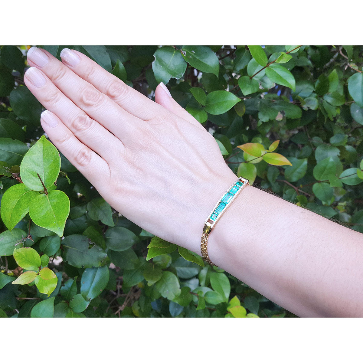 A woman&#39;s hand wearing a gold bracelet with five high quality colombian emeralds against a green leafy background.