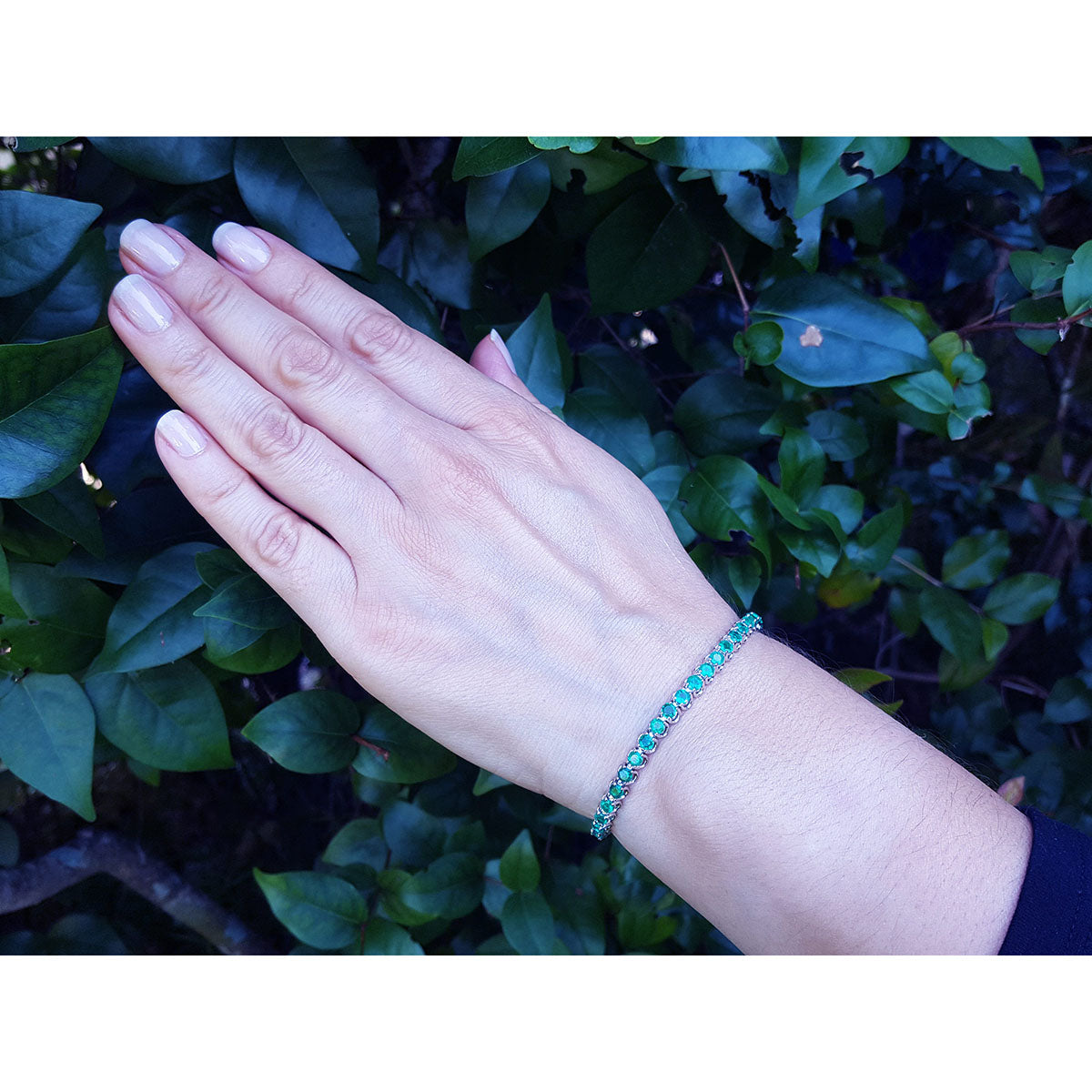 Hand wearing an emerald tennis bracelet against a green leafy background.