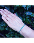 Hand wearing an emerald tennis bracelet against a green leafy background.