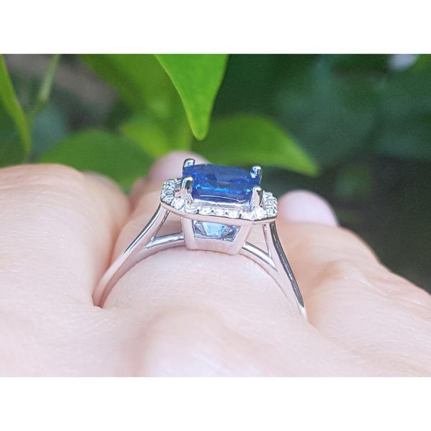 Asscher cut blue sapphire ring with halo diamonds in 18k white gold, worn on a finger.