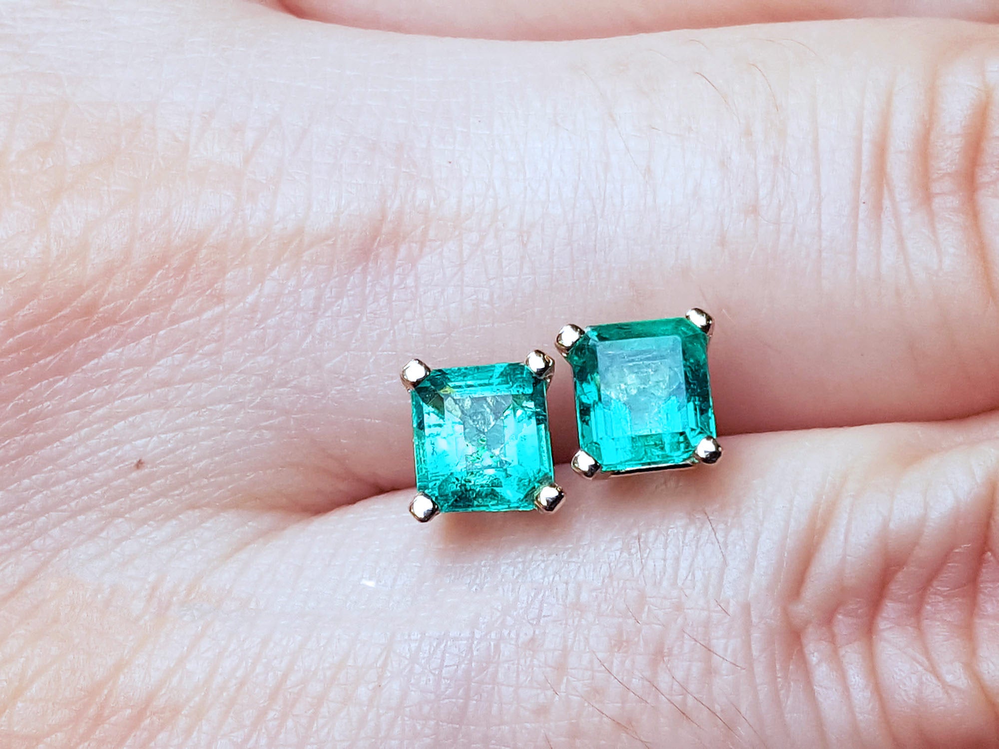  Top close view of an Asscher cut emerald stud earrings on a person&#39;s hand.