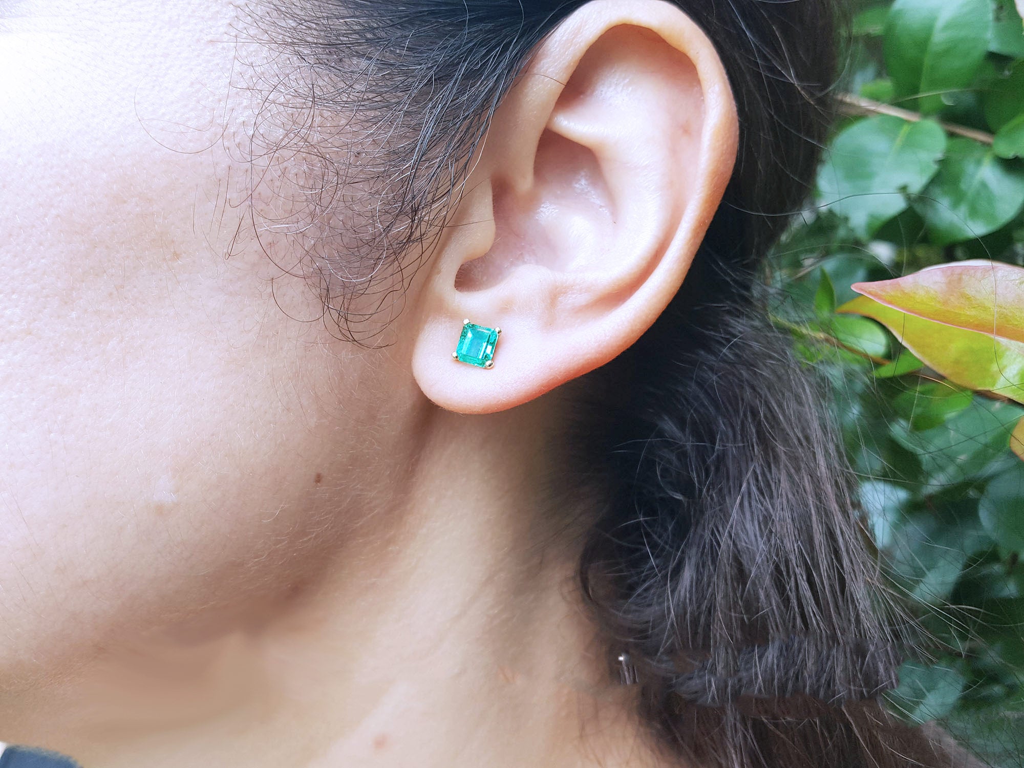 Close-up of a woman&#39;s ear wearing a 14k gold emerald stud earring with a blurred natural background