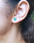 Close-up of a woman's ear wearing a 14k gold emerald stud earring with a blurred natural background