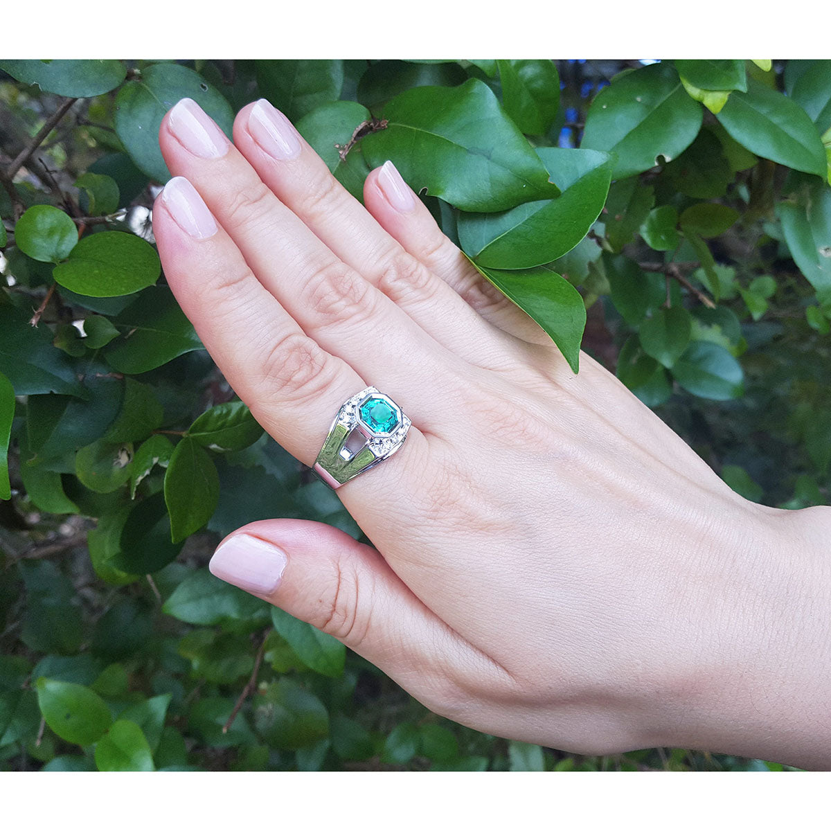 Hand wearing a white gold mens ring with a asscher cut natural colombian emerald against a green leafy background.
