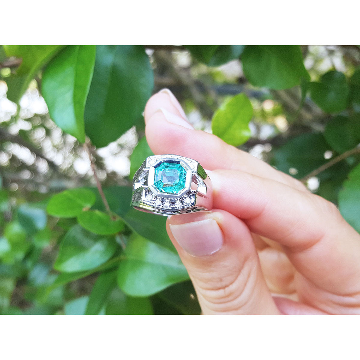 Hand holding a men&#39;s ring with a natural emerald against a blurred green leafy background,
