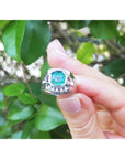 Hand holding a men's ring with a natural emerald against a blurred green leafy background,