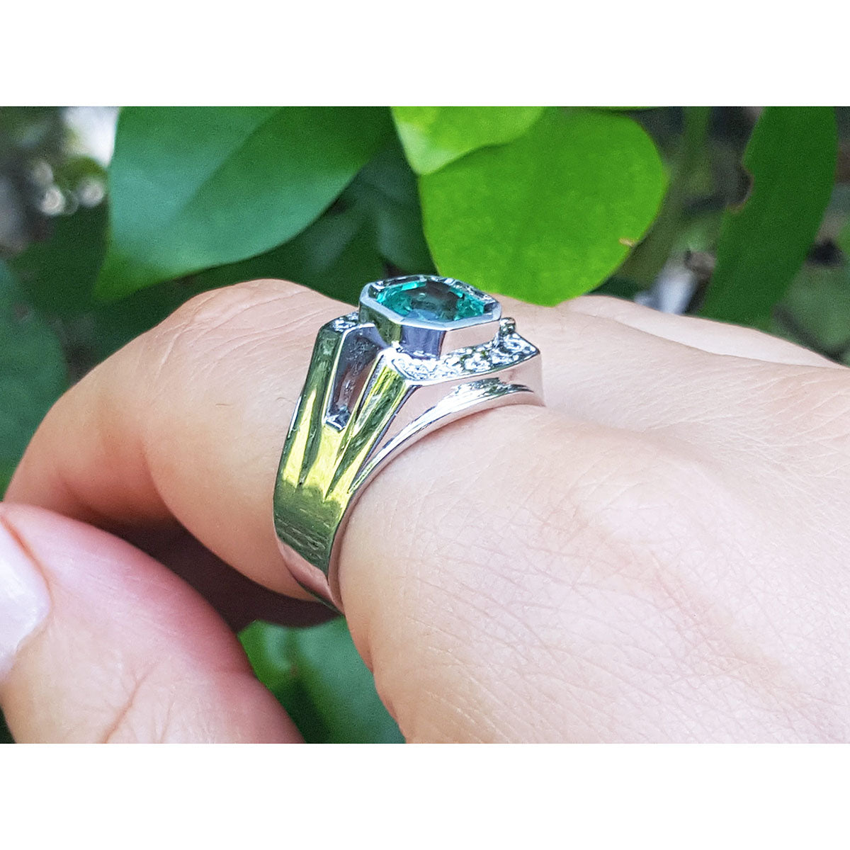 A men&#39;s emerald ring underside view on a finger against a green leafy background.
