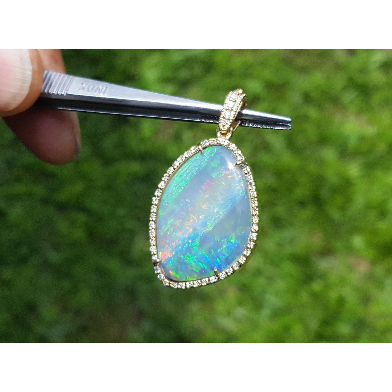 Opal pendant held by tweezers with a blurred green background.
