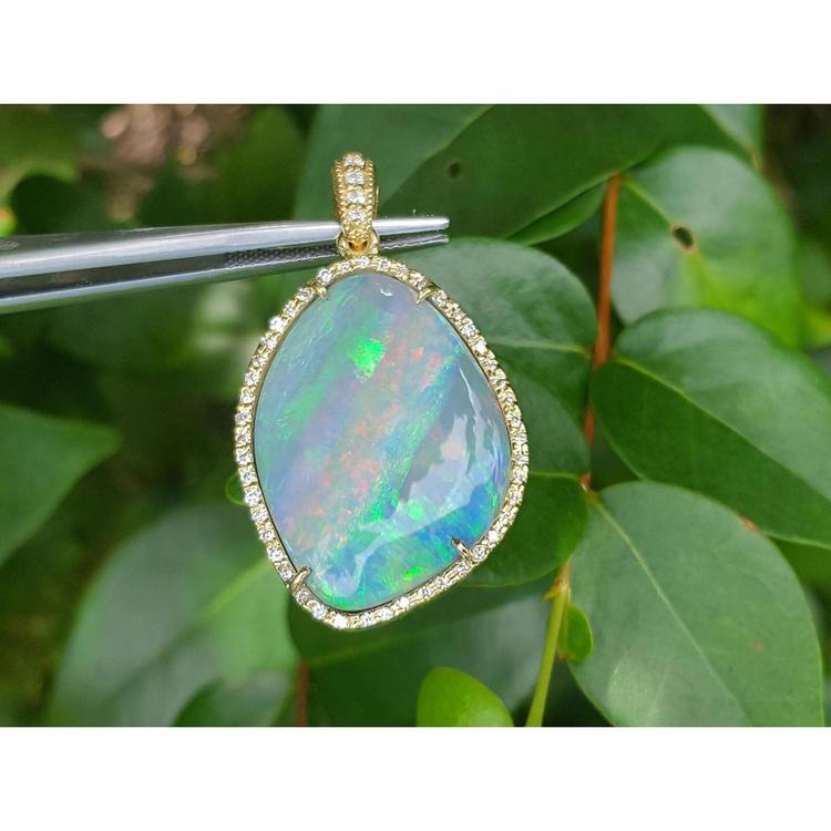 Opal pendant with gold setting held by tweezers against a green leafy background.