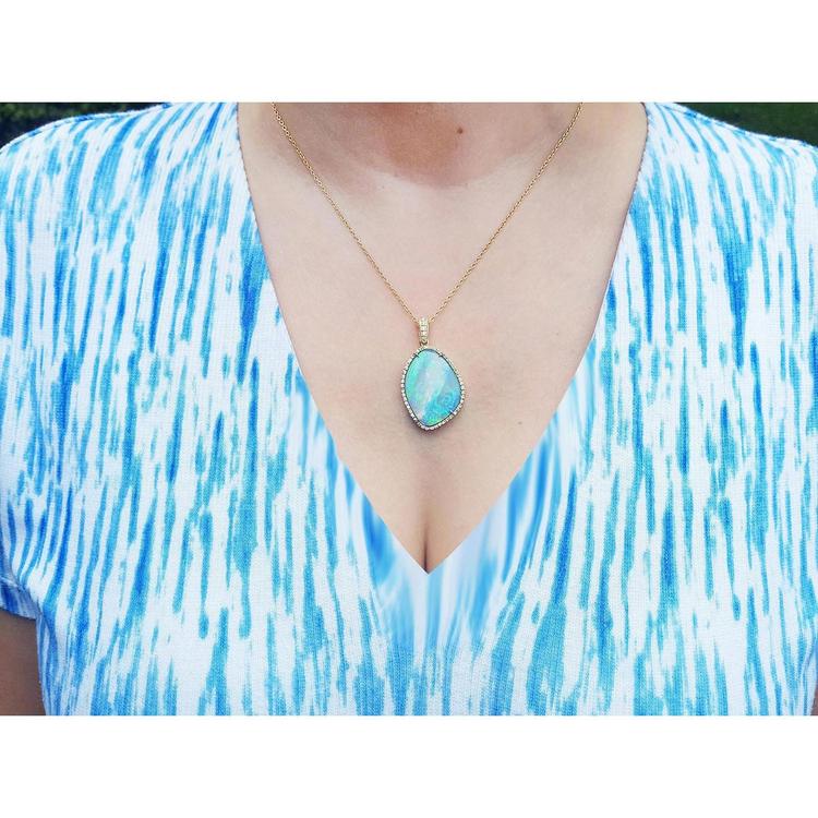 A woman wearing a blue and white patterned top with a gold necklace featuring a natural opal pendant.
