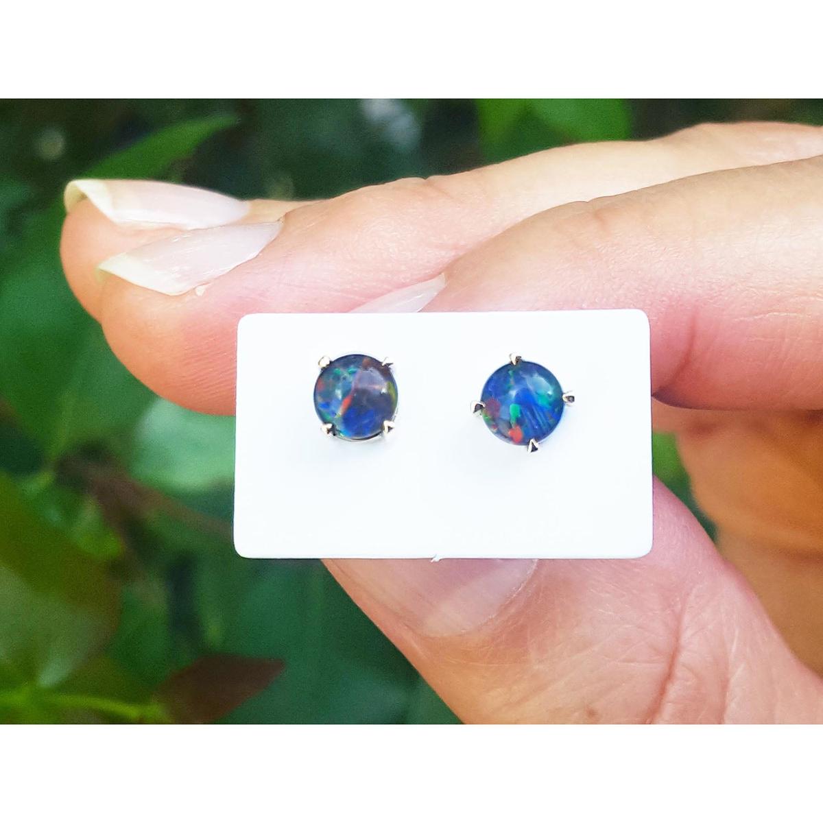 Opal stud earrings on a white card held between fingers with a blurred green background.