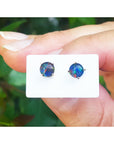 Opal stud earrings on a white card held between fingers with a blurred green background.