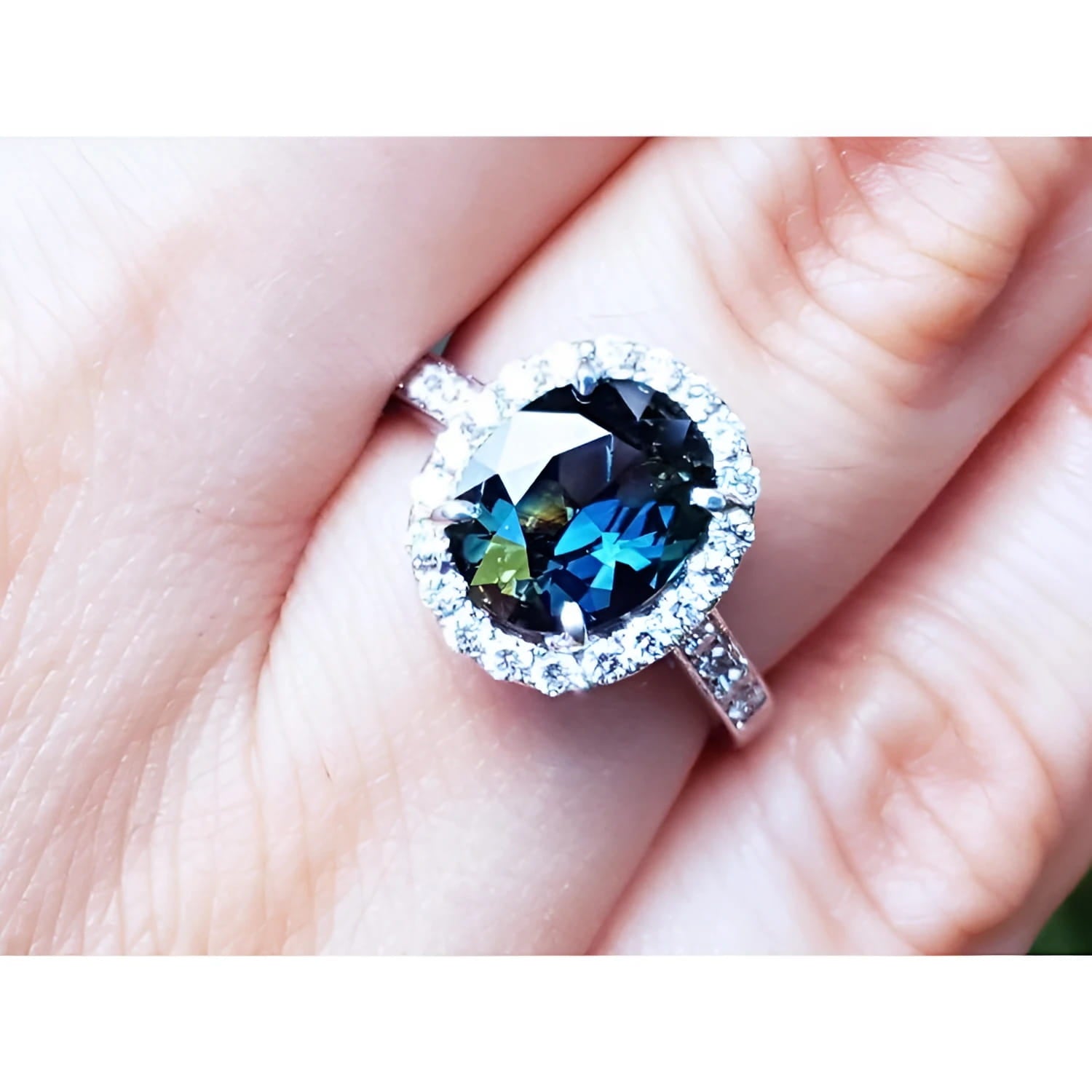 An oval-cut Australian sapphire ring for women, featuring a sparkling bluish-green hue, surrounded by a halo of diamonds set in 18K white gold, worn on a finger.