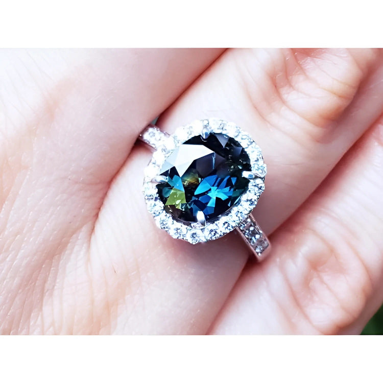 An oval-cut Australian sapphire ring for women, featuring a sparkling bluish-green hue, surrounded by a halo of diamonds set in 18K white gold, worn on a finger.