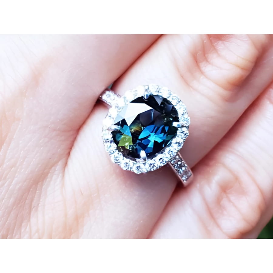 An oval-cut Australian sapphire ring for women, featuring a sparkling bluish-green hue, surrounded by a halo of diamonds set in 18K white gold, worn on a finger.