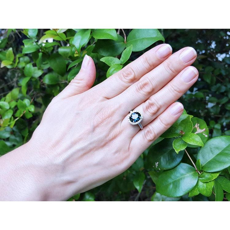 Oval bluish-green Australian sapphire ring with diamond halo, set in 18K white gold on a woman's hand.