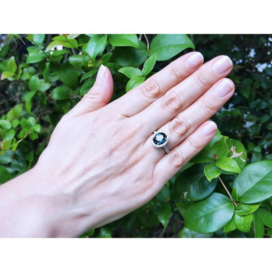 Oval bluish-green Australian sapphire ring with diamond halo, set in 18K white gold on a woman's hand.