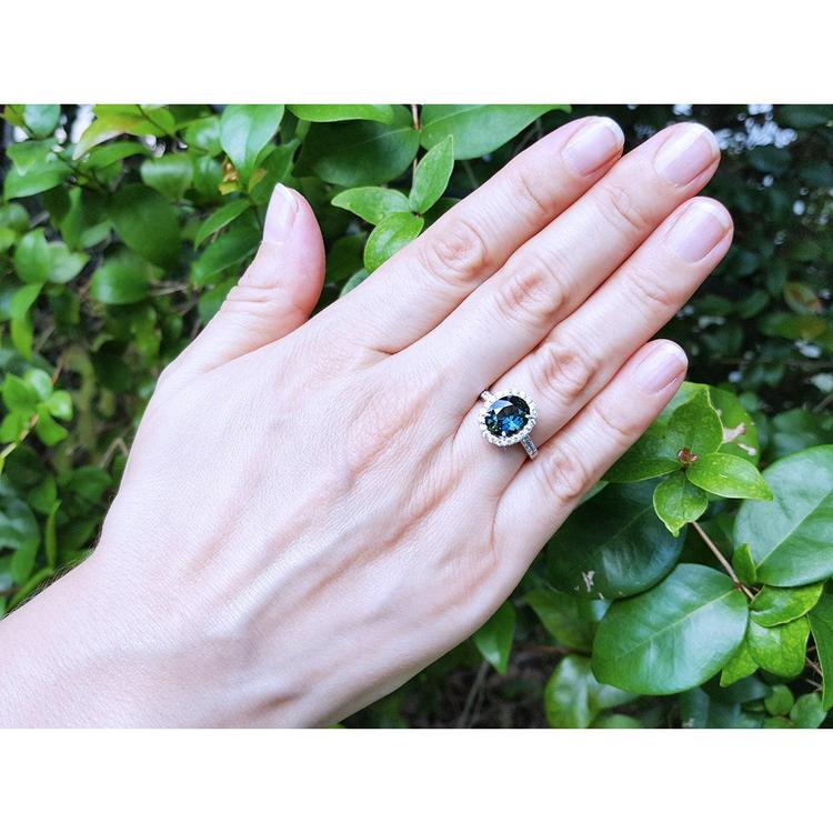 Oval bluish-green Australian sapphire ring with diamond halo, set in 18K white gold on a woman's hand.