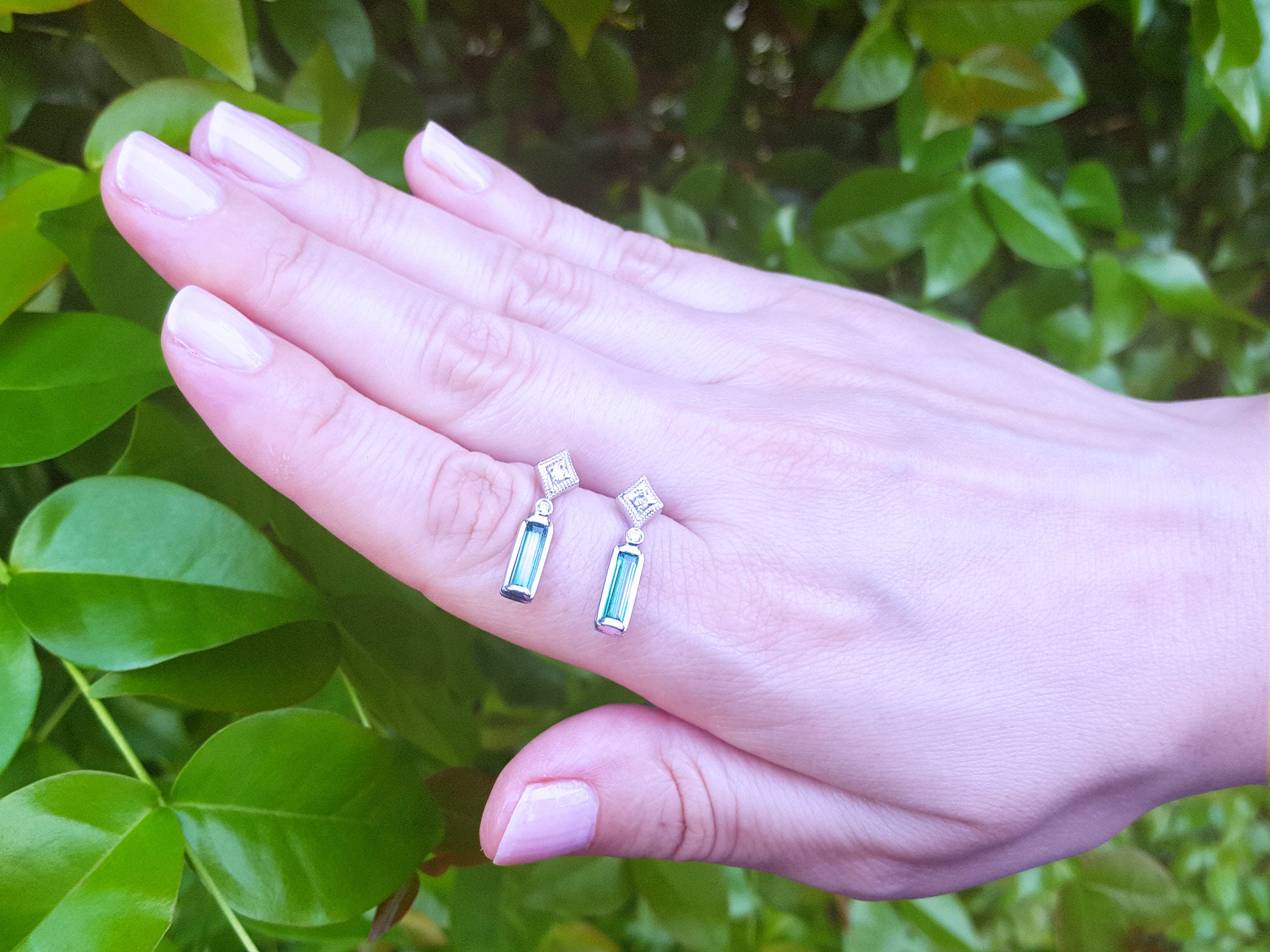Hand displaying two emerald earrings with diamond and white gold against a green leafy background
