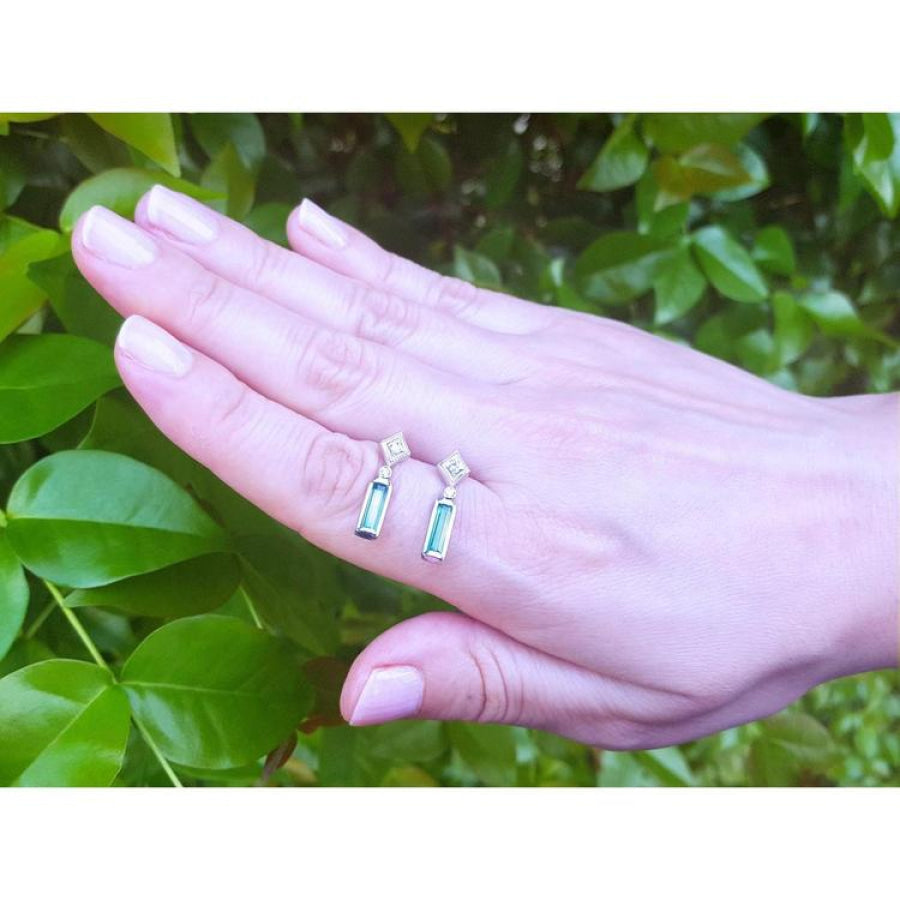 14K white gold emerald baguette cut earrings with diamond studs, worn on a hand against green foliage.