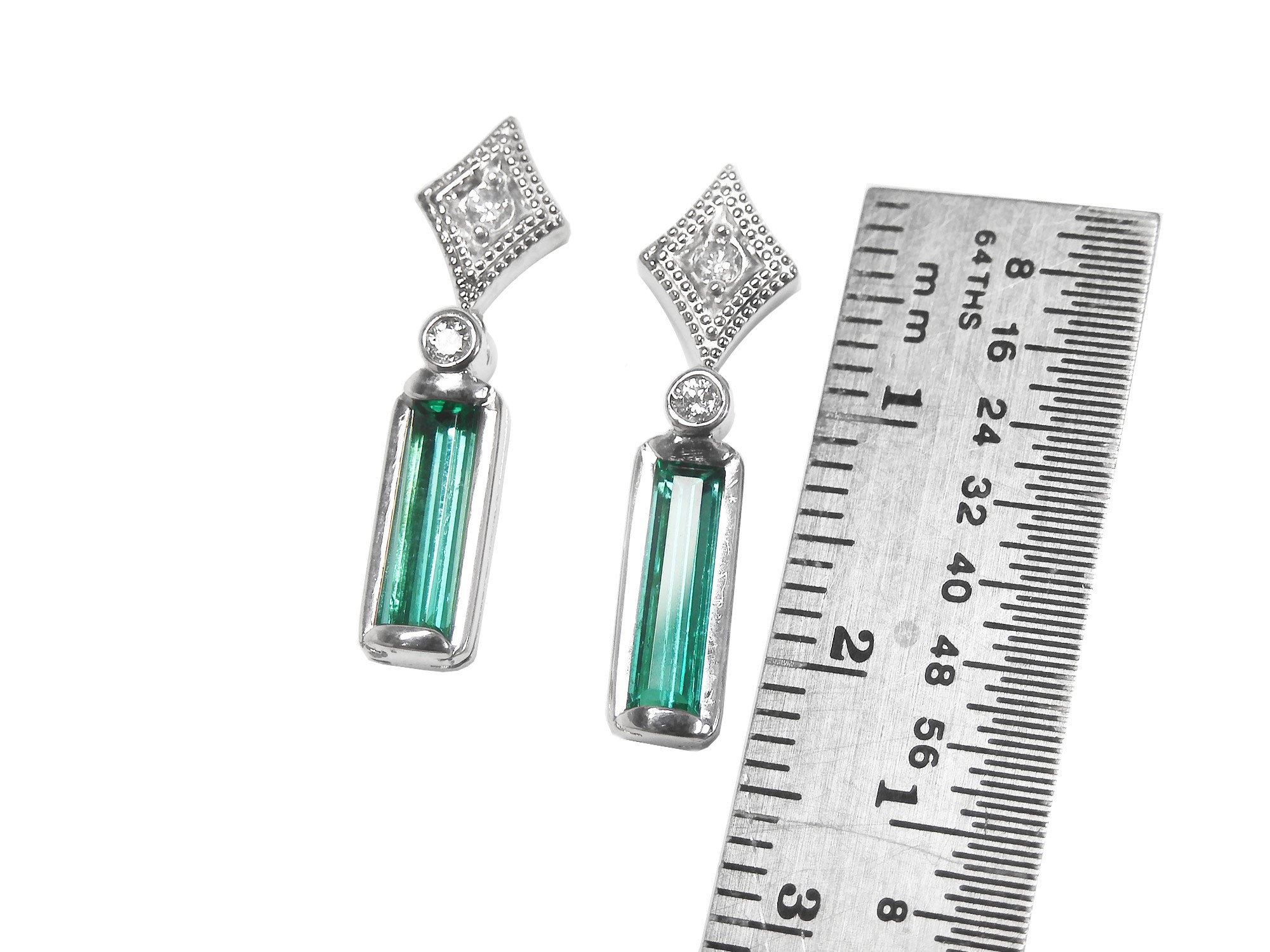 Pair of earrings with natural emeralds and diamond tops next to a ruler for scale on a white background