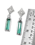 Pair of earrings with natural emeralds and diamond tops next to a ruler for scale on a white background