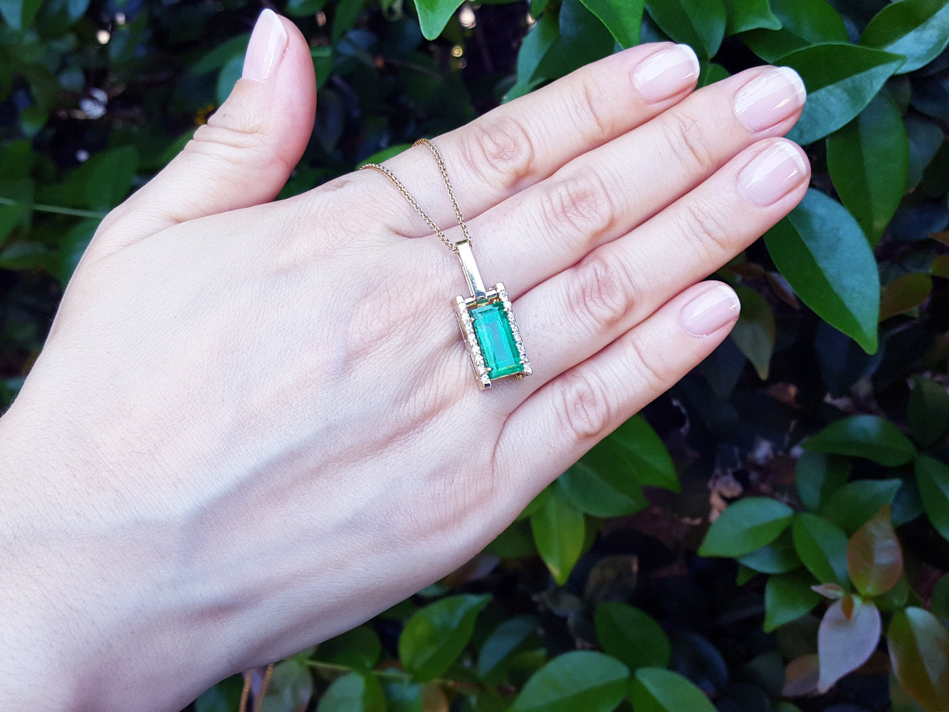 Hand displaying a gold pendant necklace with a natural emerald against a leafy background.