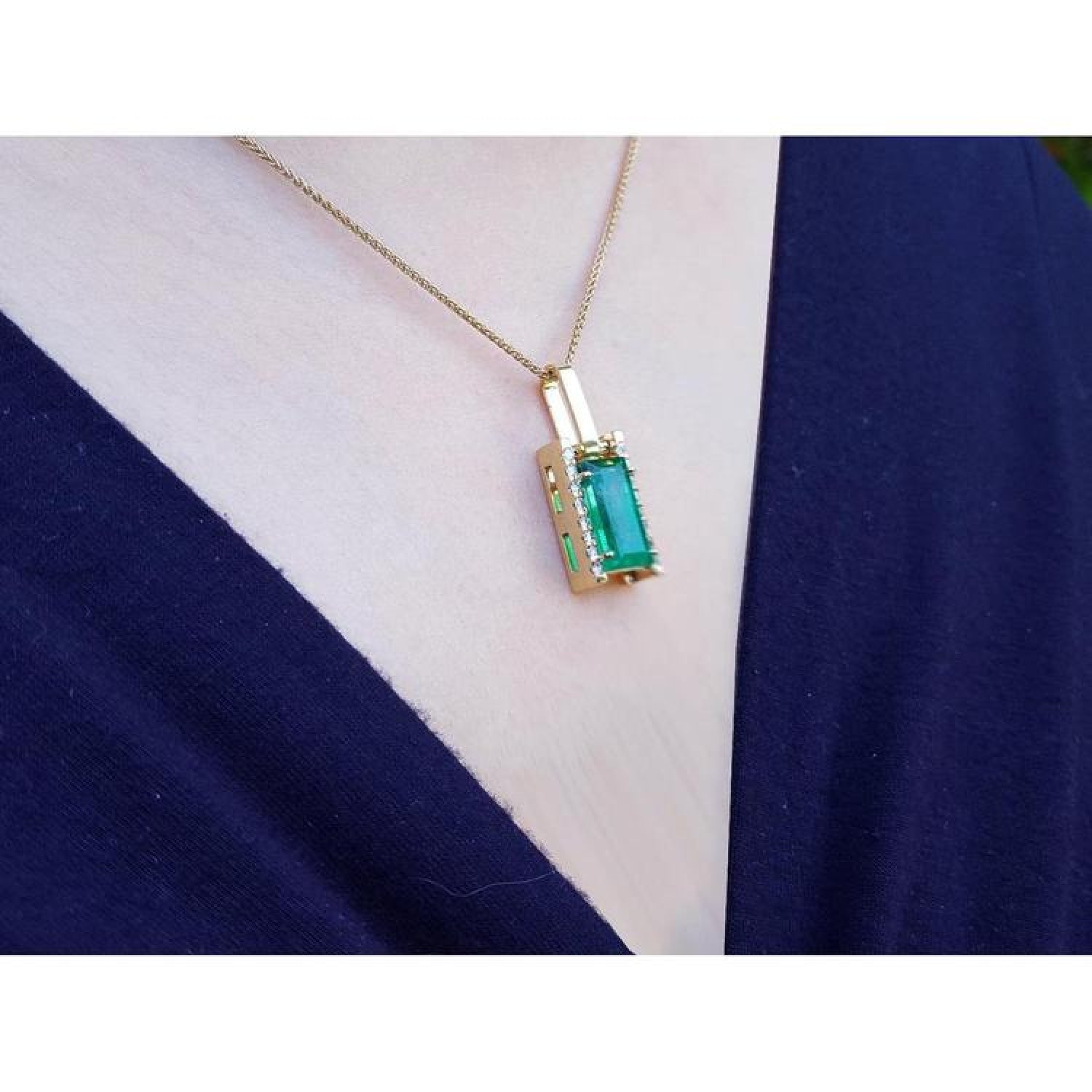 Baguette cut emerald pendant in 18K yellow gold with diamond accents, worn on a dark blue top.