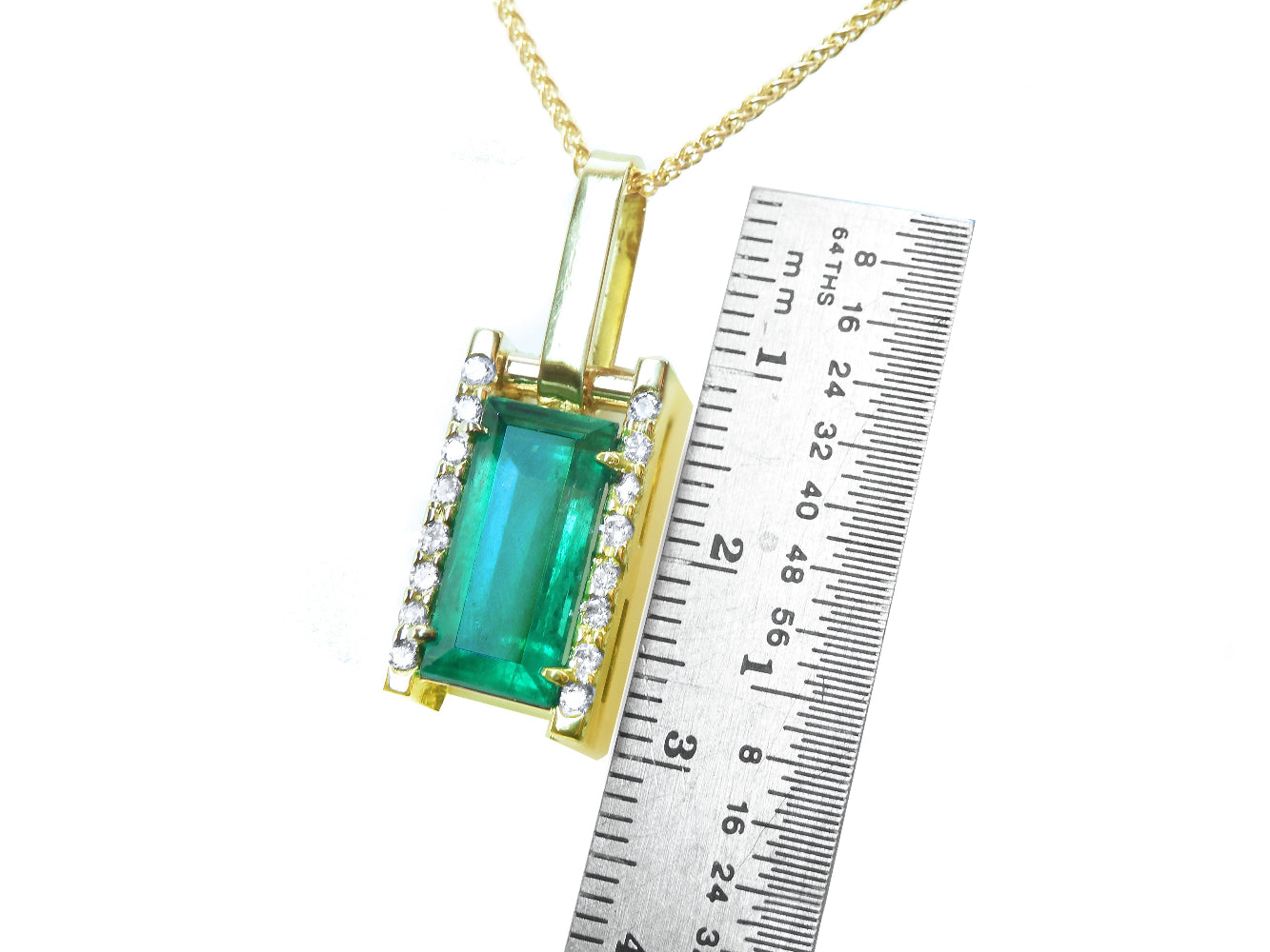 Gold pendant with a baguette cut emerald next to a ruler for scale on a white background.