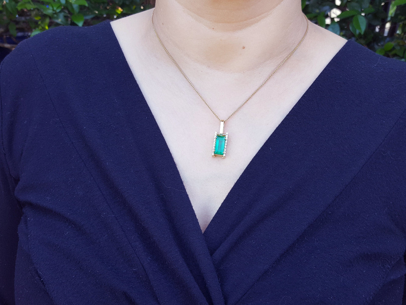 A woman wearing a navy blue outfit with a gold necklace featuring a baguette cut emerald pendant.