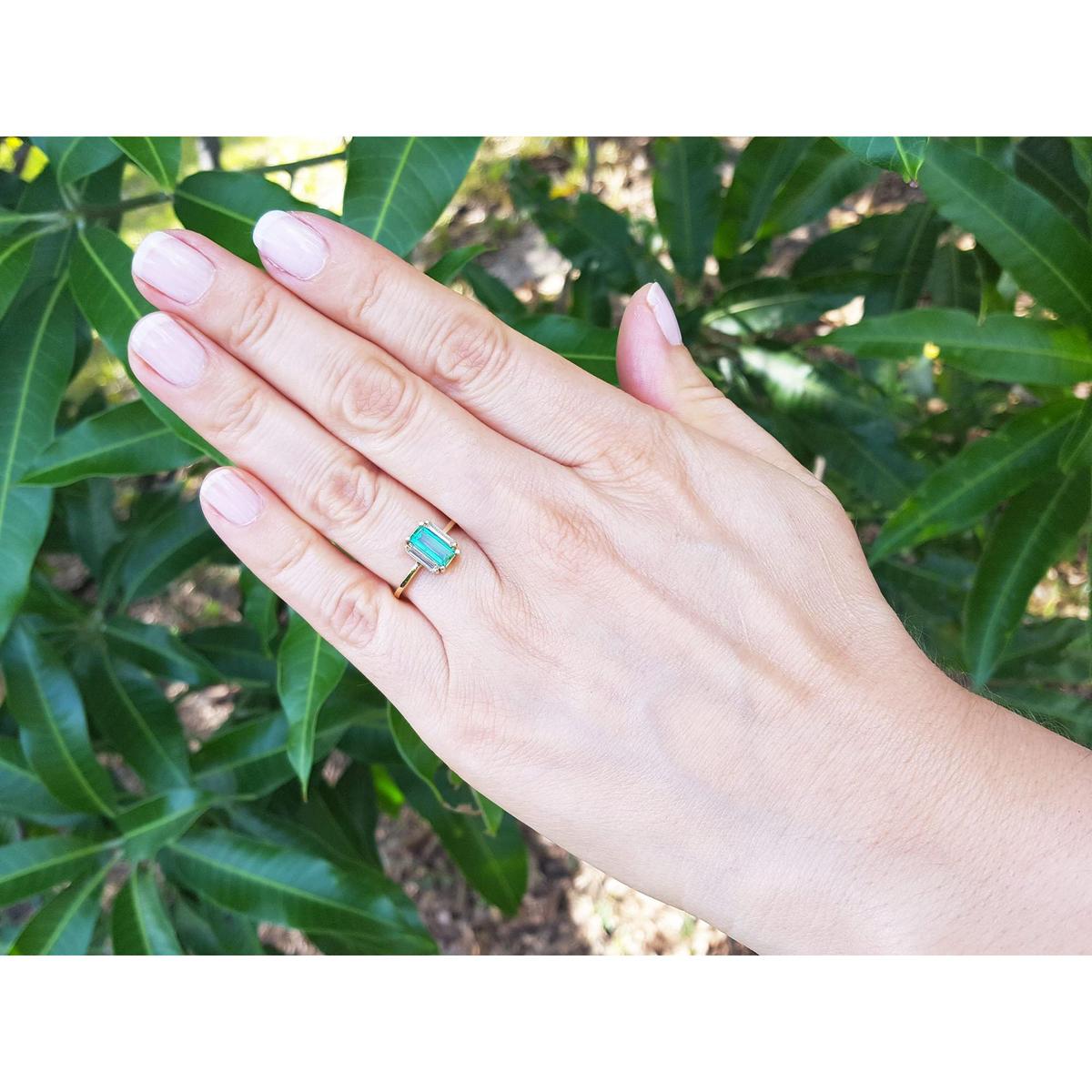 Hand wearing a baguette emerald and side diamonds ring with green leaves in the background.