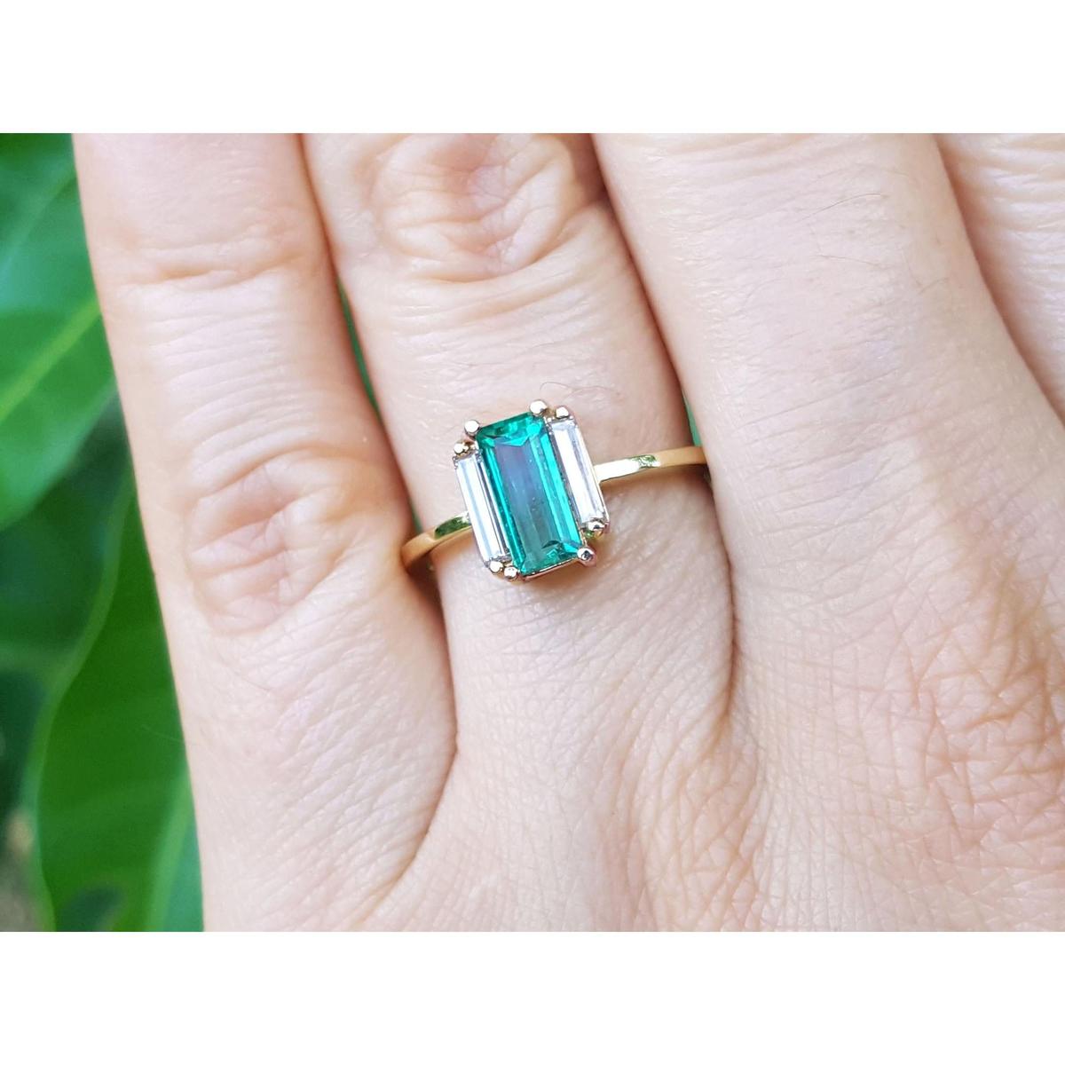 Emerald and side straight baguettes in 18k gold ring on a person's finger against a blurred natural background.