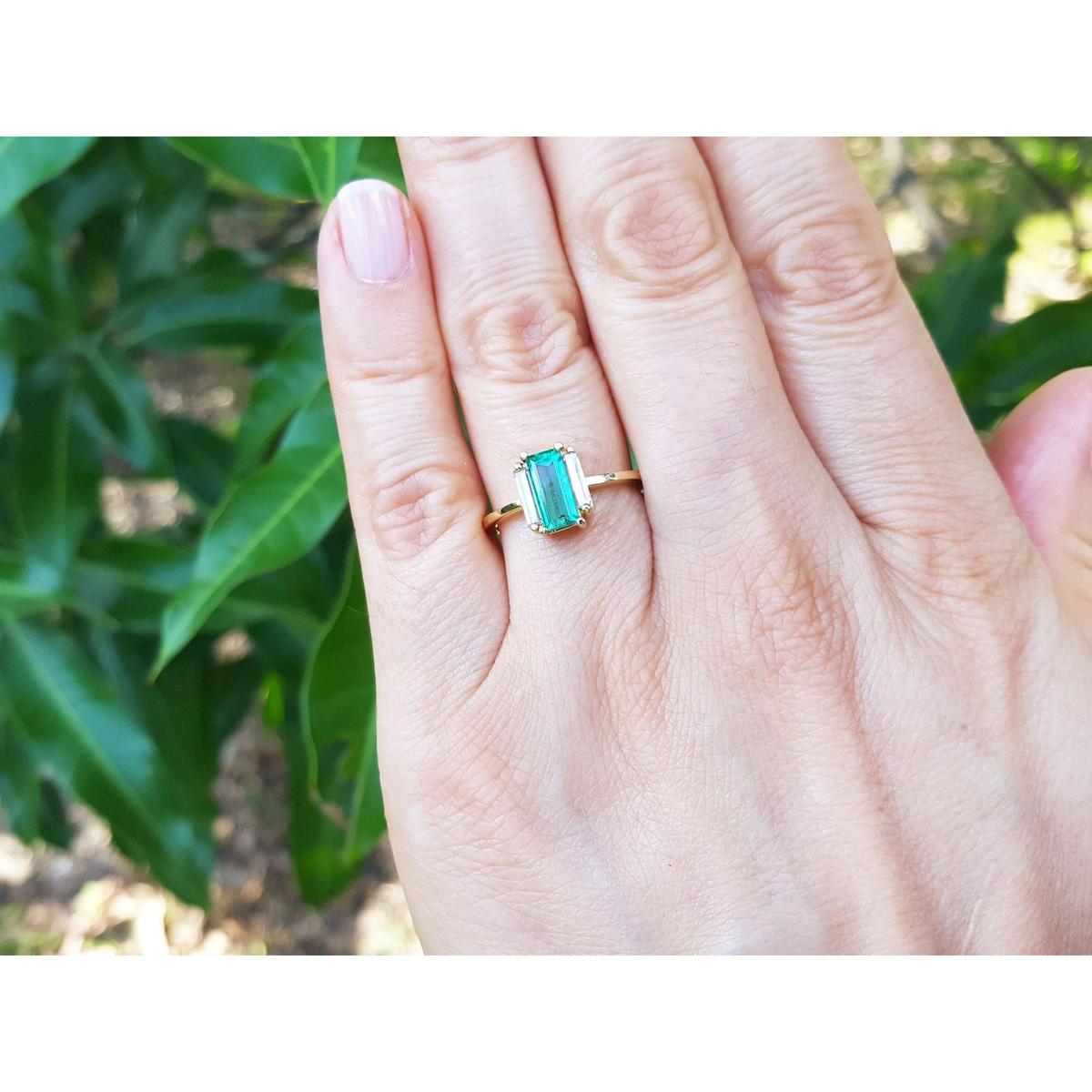 Hand wearing a ring with emerald baguette cut and diamonds each side against a leafy background.