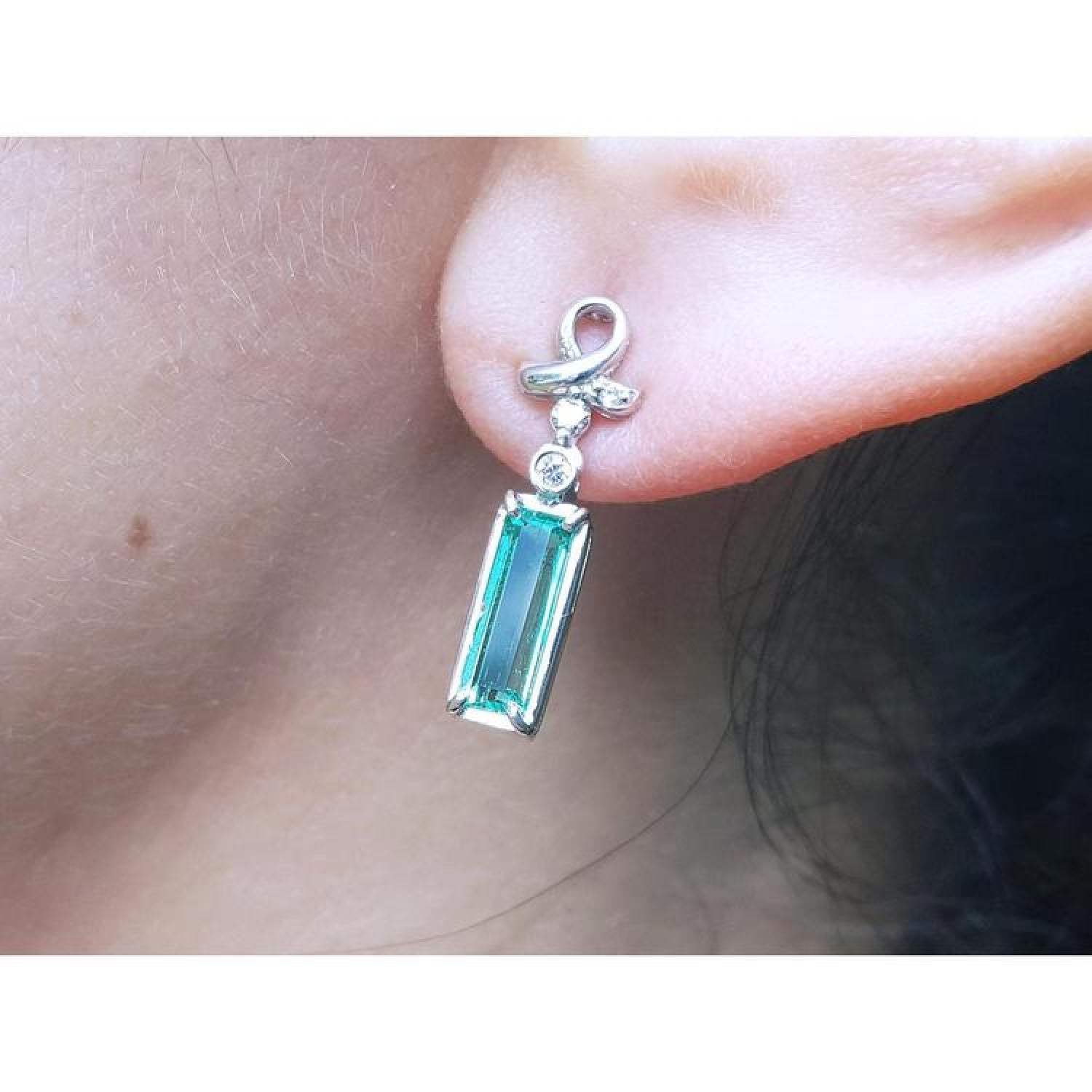 Emerald-cut natural emerald earrings in 14k white gold with ribbon design and accent diamonds.
