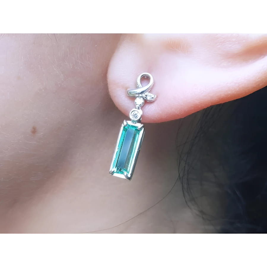 Emerald-cut natural emerald earrings in 14k white gold with ribbon design and accent diamonds.