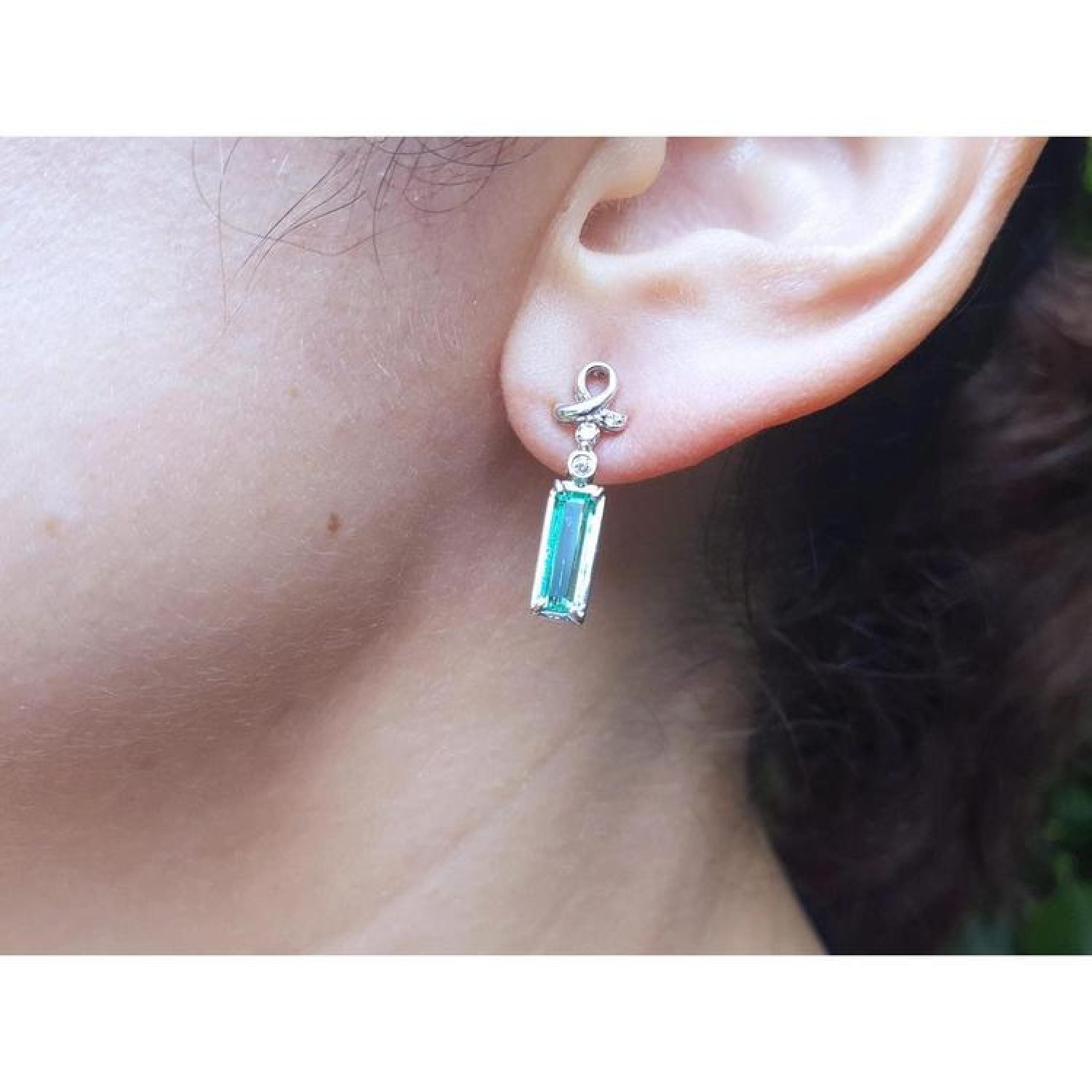 Emerald-cut natural emerald earrings in 14k white gold with diamond accents, dangling from a ribbon design.