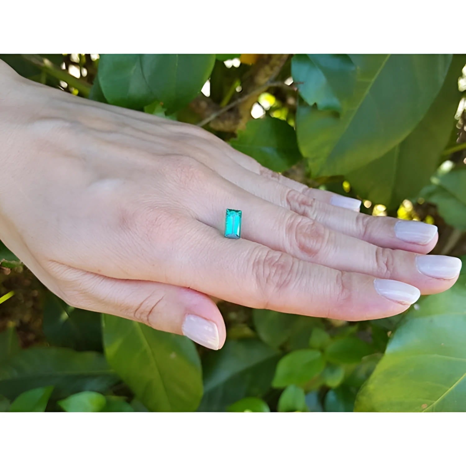 1.62 ct. baguette-cut loose natural emerald, vibrant green, held on a hand against foliage.